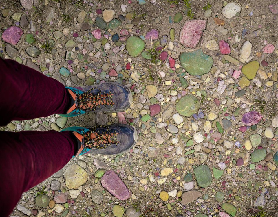 A photo of colourful rocks dotting a trail, taken from the perspective of someone holding a camera and pointing it at their feet. The colours of the rocks include a jade green, several purple shades, a variety of pink shades, yellow, and red. The photo also includes purple hiking boots with blue and orange accents. There are also red pants.