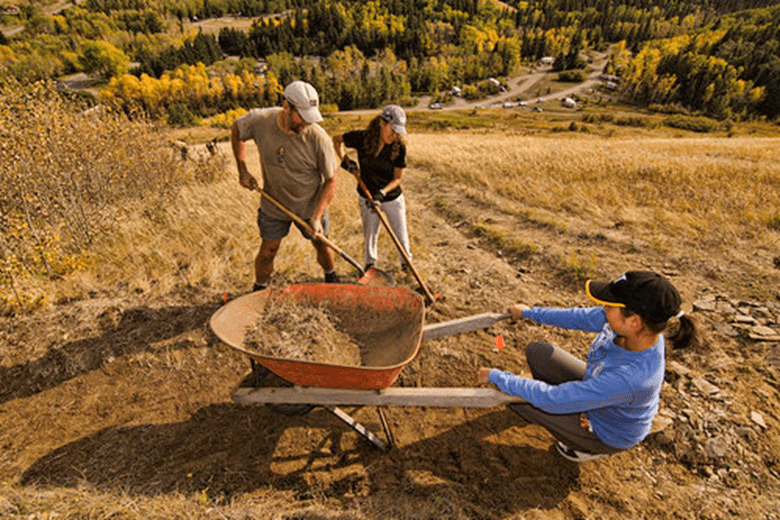 Three Alberta Parks volunteers at work. One is holding a wheelbarrow in place while the other two shovel dirt into the wheelbarrow.