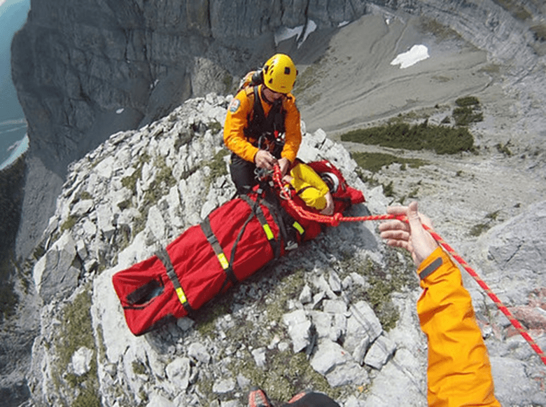 A photo taken from the perspective of a rescuer. In the photo, you can see another rescuer securing a person in a heli-bag in a remote location on top of a mountain.