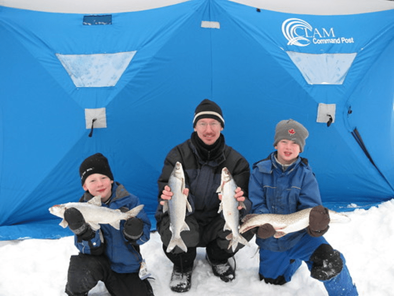 Two children and an adult hold up 4 fish in front of a blue ice fishing tent