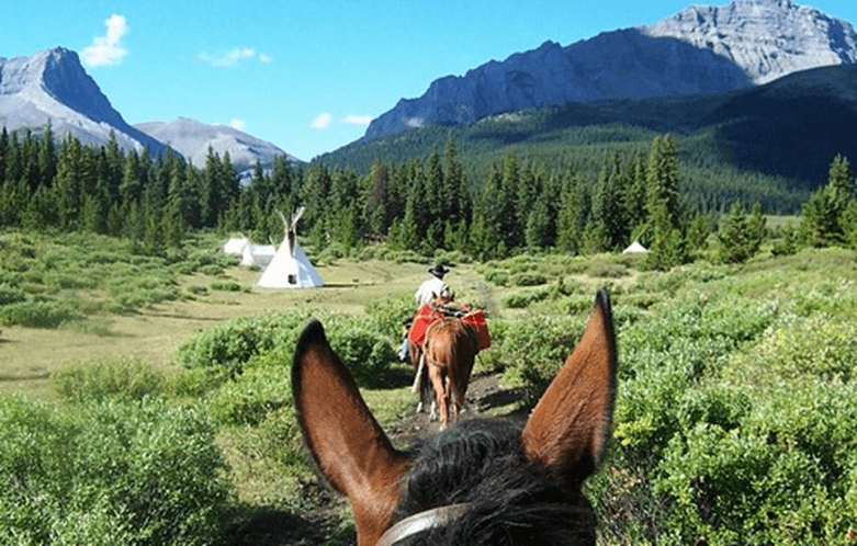 A photo taken from the back of a horse as if you were facing forward. In front of you, there is another horse packing supplies and another person. To the left of this, you can see a white teepee. In the background, you can see a pine forest and mountains with steep cliffs.
