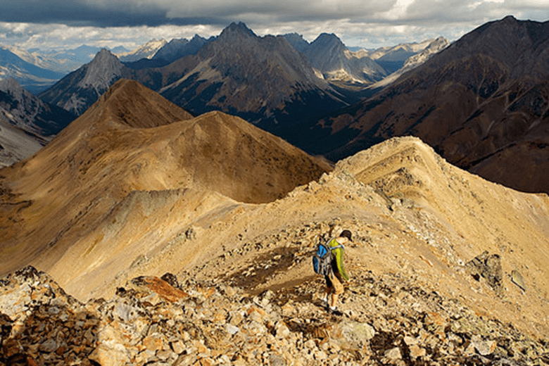 A photo of a hiker on Pocaterra Ridge. The soil on the mountain seems tinted orange. In the background, you can see various mountains.